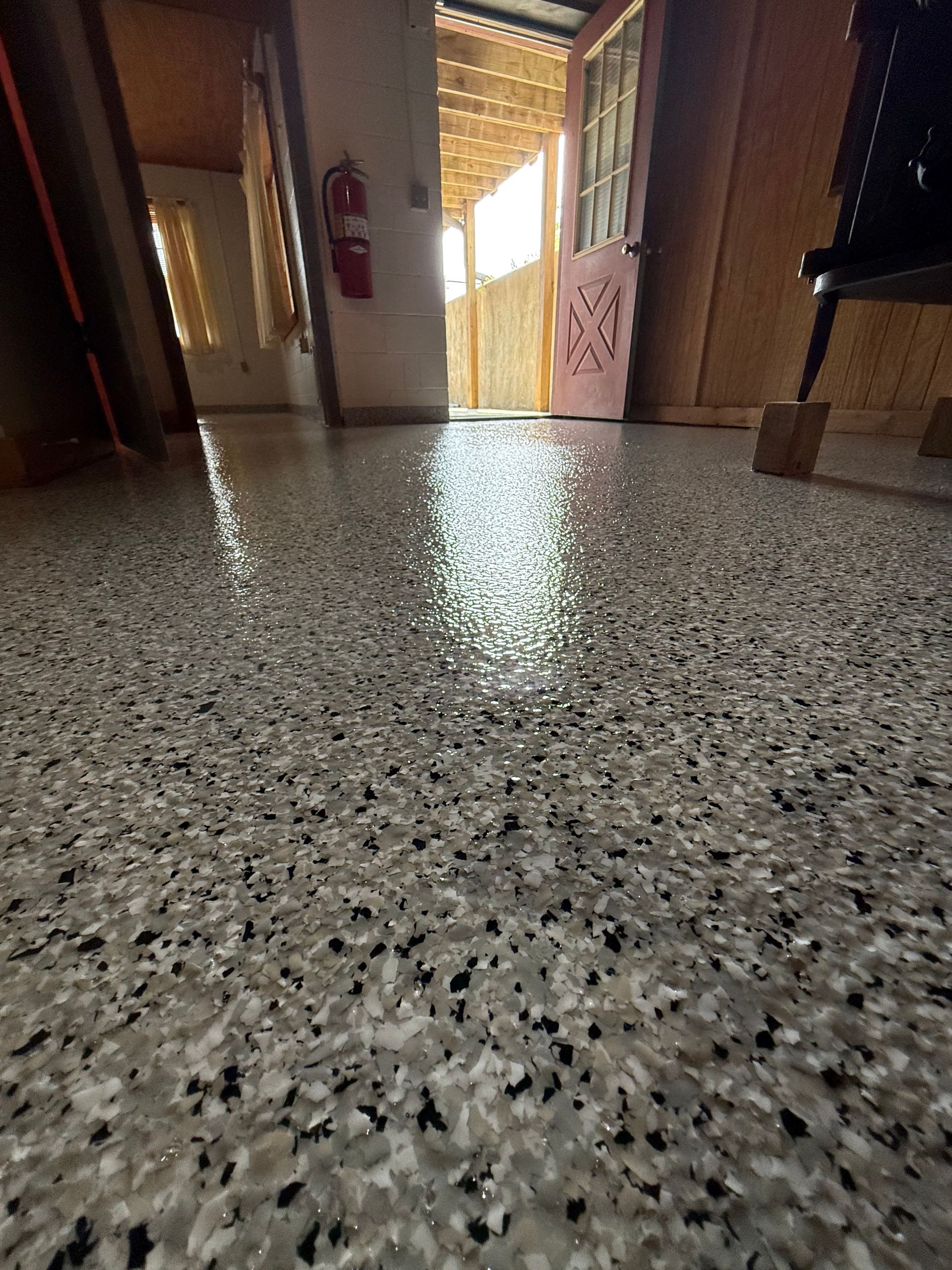 Low-angle view of a terrazzo hallway floor leading to a bright doorway, with doors and a fire extinguisher on the walls.
