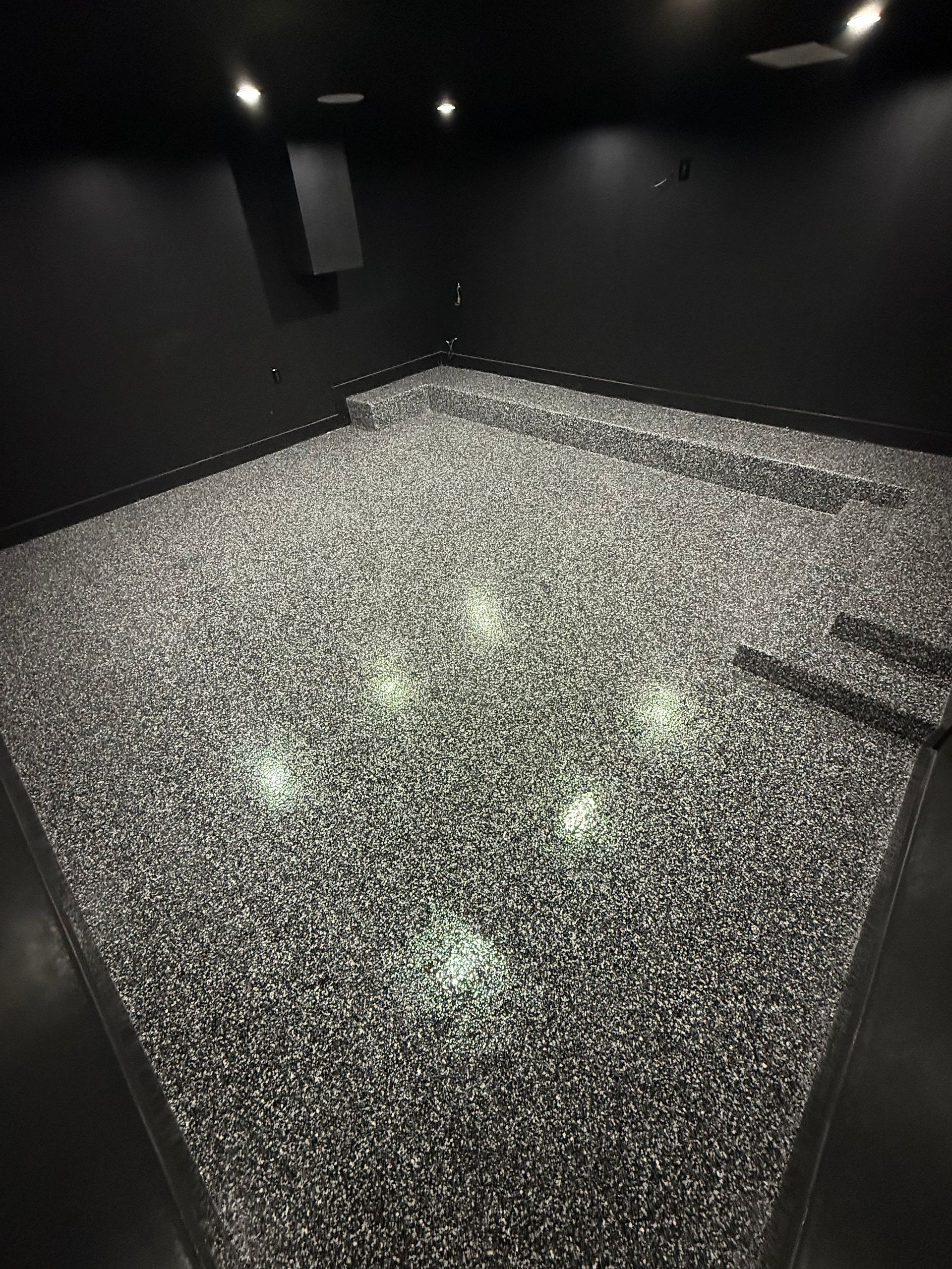 Empty dark room with black walls and a speckled gray floor under ceiling lights.