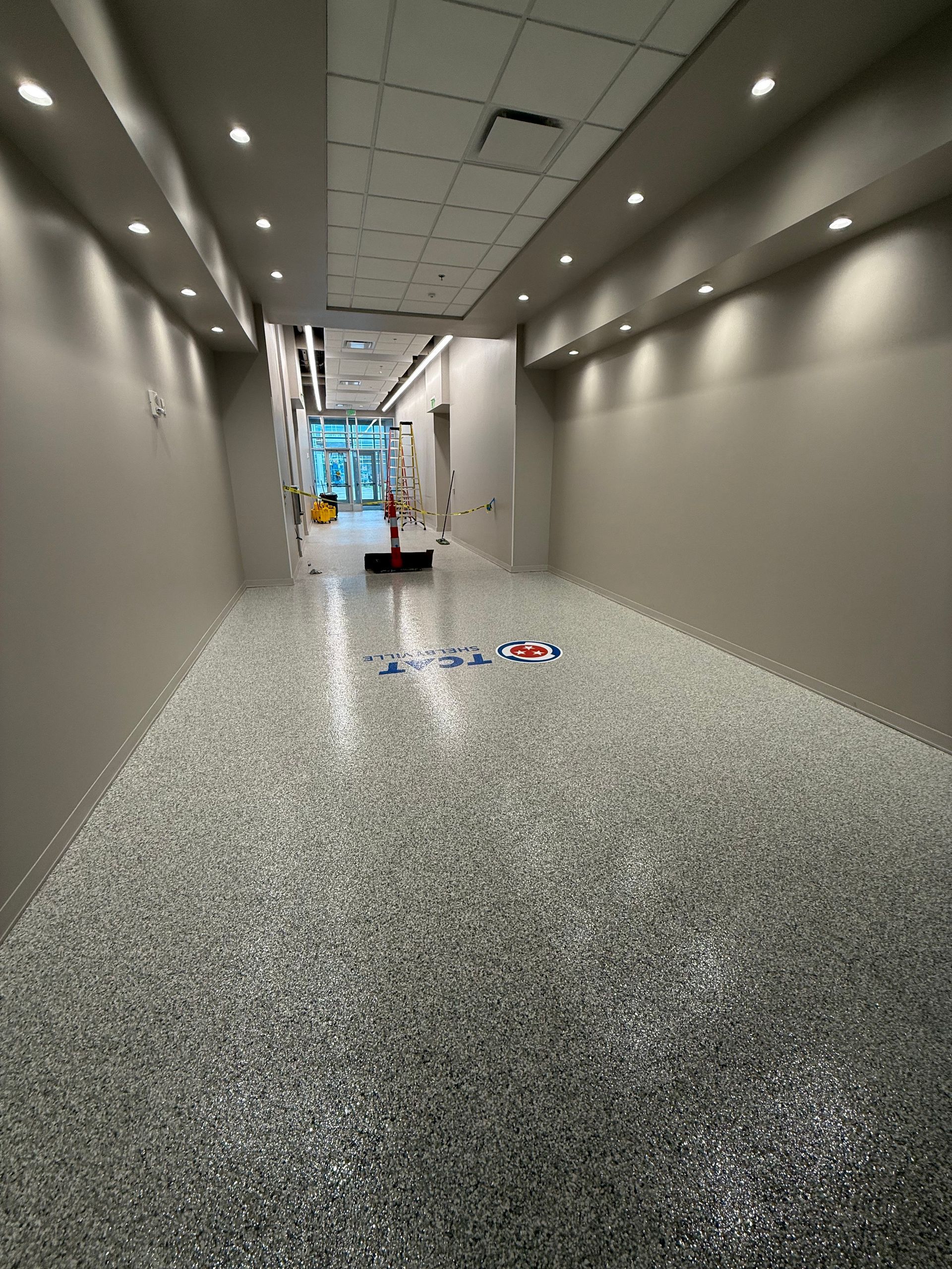 Long hallway with gray walls, speckled floor, recessed lights, and cleaning supplies near the far end.