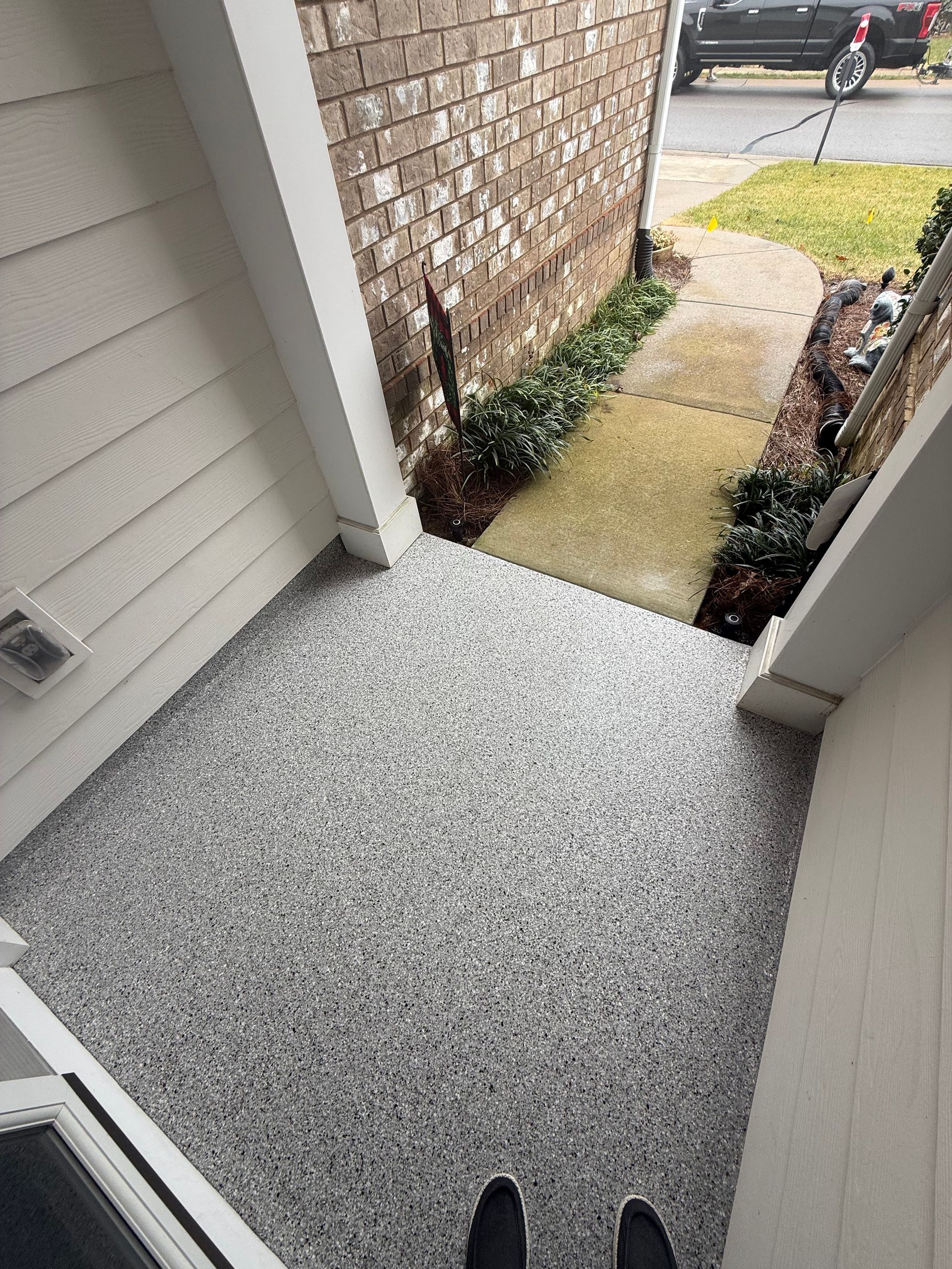 Freshly coated gray patio walkway between beige siding and a brick wall, leading to a front yard path.