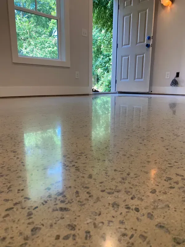 Empty room with polished speckled concrete floor, window, and open front door showing greenery outside