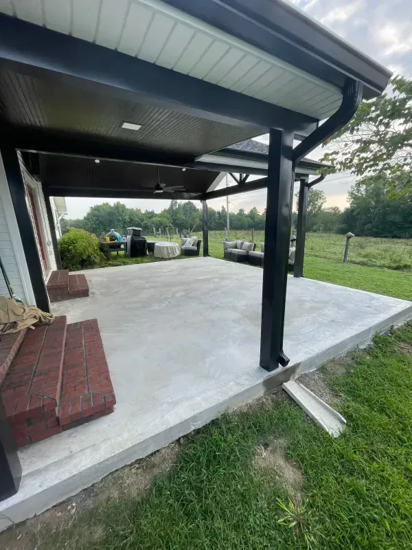 Covered concrete patio with black support posts beside a grassy yard and brick steps
