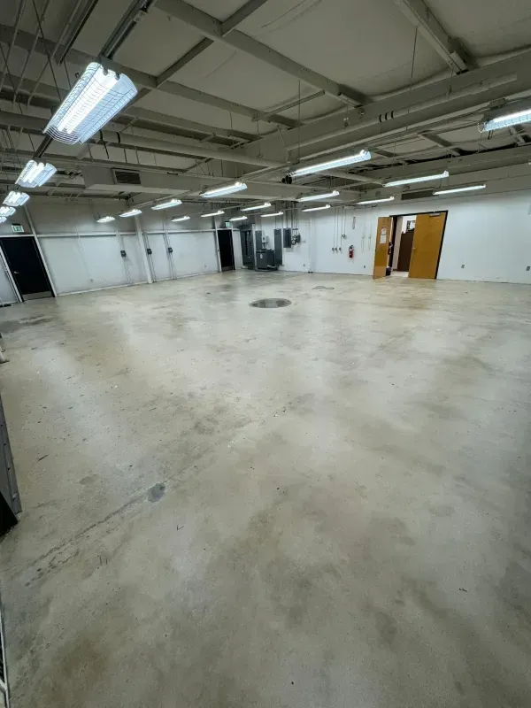 Empty industrial room with concrete floor, white walls, exposed ceiling, and fluorescent lights