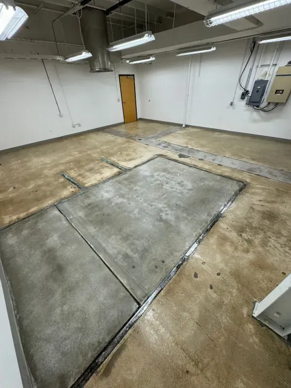 Empty industrial room with a large rectangular concrete patch in the floor and white walls under fluorescent lights