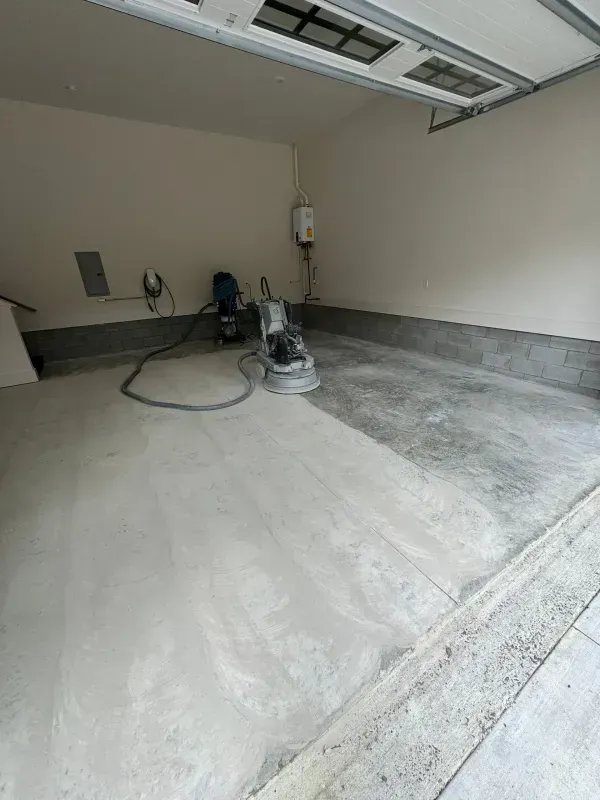 Empty garage with concrete floor being sanded by a floor grinder, dust-covered surface and open garage door.