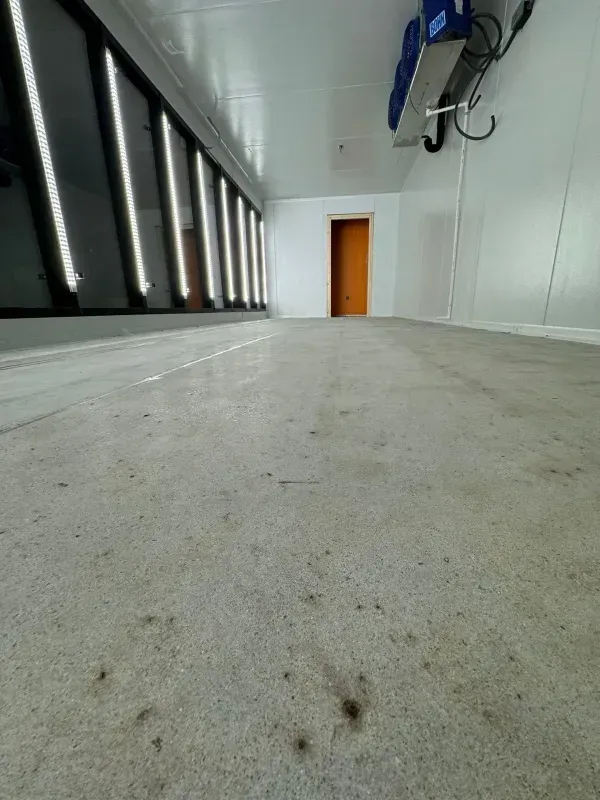 Long industrial hallway with concrete floor, white walls, and an orange door at the end