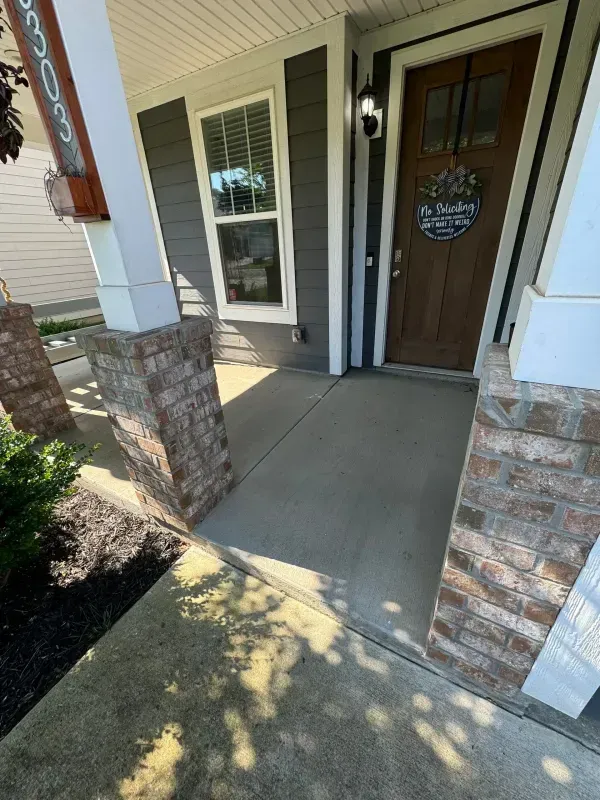 Front porch with concrete walkway, stone columns, and a wooden front door beside a window.