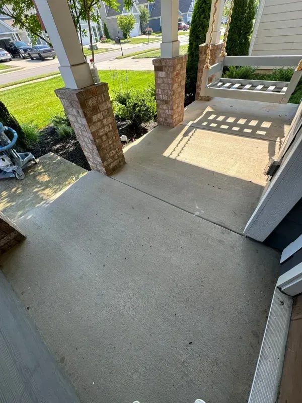 Front porch concrete walkway with brick columns, white railings, and a bright sunlit sidewalk beyond.
