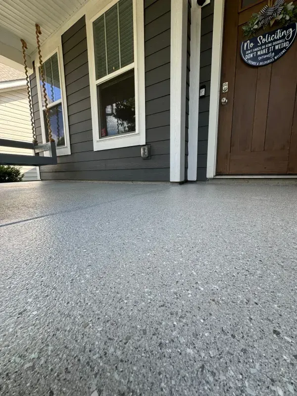 Low-angle view of a house porch with gray siding, white trim, a brown door, and a speckled concrete floor