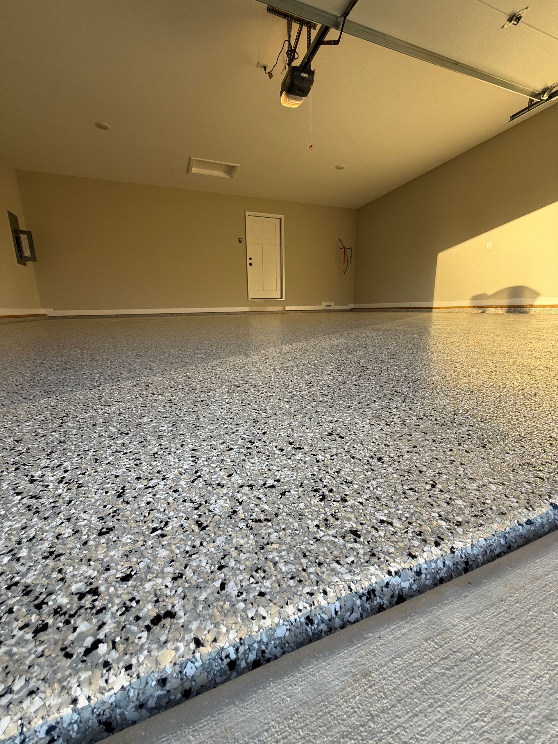 Empty garage with speckled epoxy floor and closed white door, lit by sunlight.