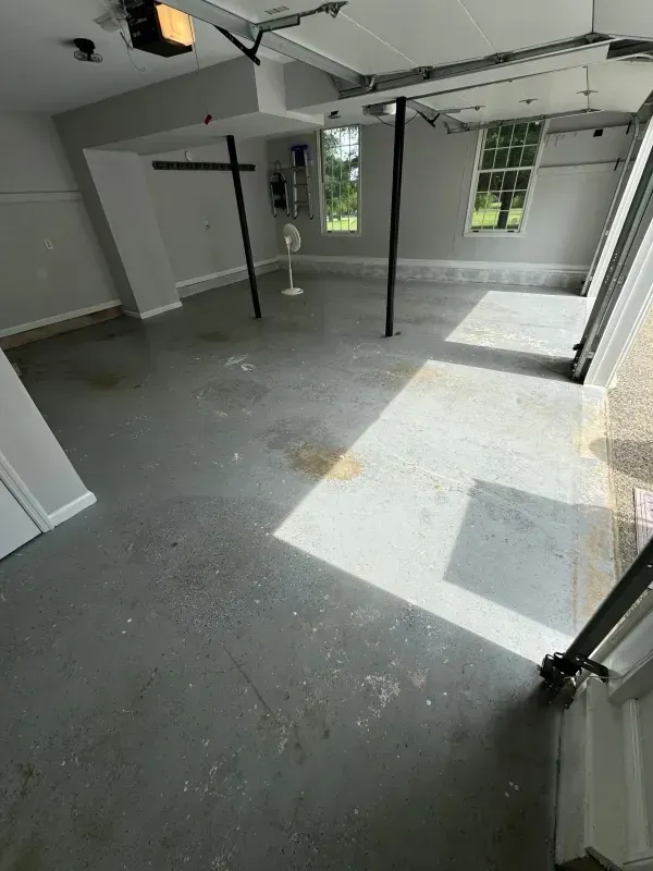 Empty unfinished room with gray floor, exposed beams, and sunlight from a window.