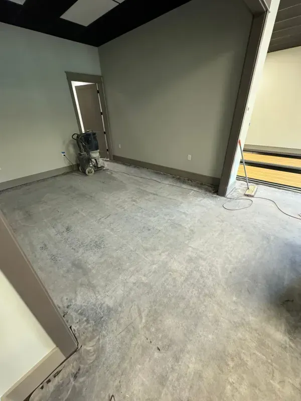 Empty gray room with unfinished concrete floor, open doorway, and construction tools near the wall