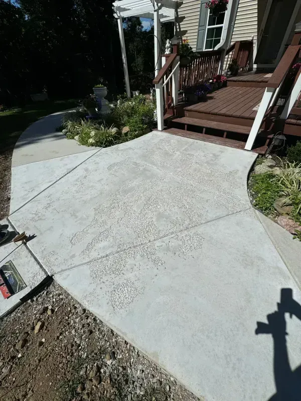 Fresh concrete walkway beside a house porch with steps, railing, and garden beds in sunlight