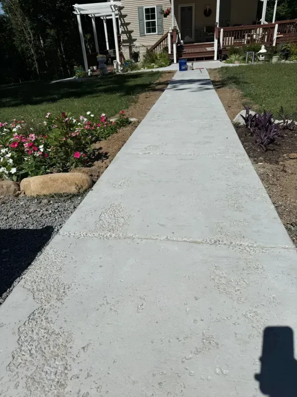 Concrete walkway leading to a house, bordered by flower beds and garden stones.