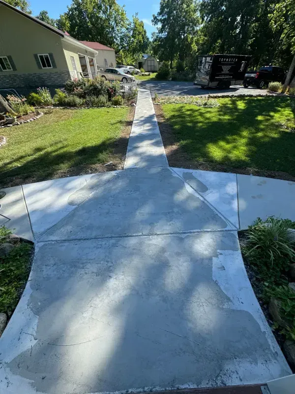 Concrete sidewalk leading to a house, bordered by green lawn and trees on a sunny day