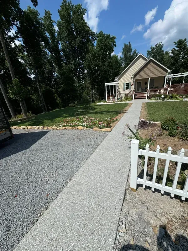 Gravel driveway and walkway leading to a beige house with white trim, lawn, and a small white fence in front.