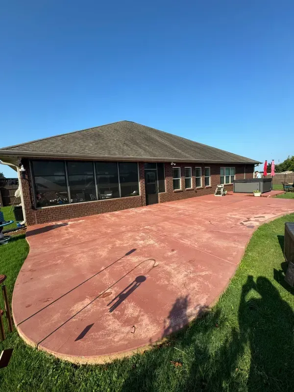 Backyard patio with a brick house, red concrete slab, and green lawn under a clear blue sky