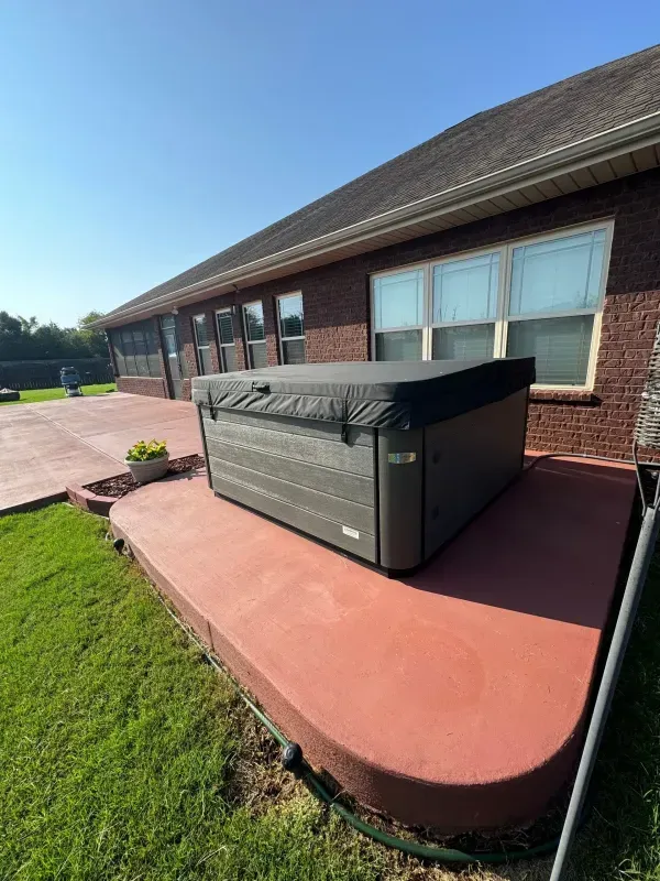 Hot tub on a red patio beside a brick building with windows and a grassy lawn.