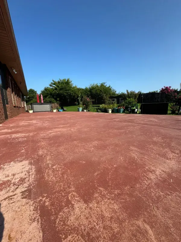 Outdoor patio with a red concrete surface, brick wall on the left, and green trees under a blue sky.