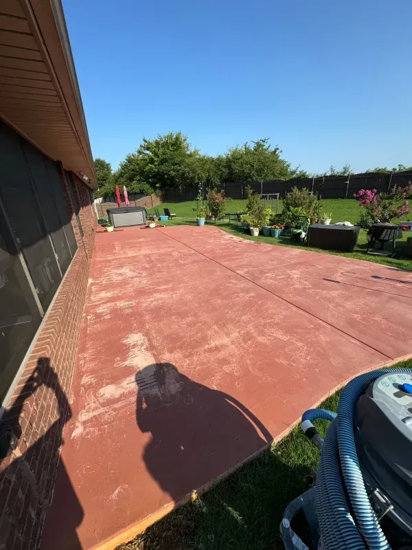 Backyard patio with red concrete, garden beds, and a house wall under a blue sky