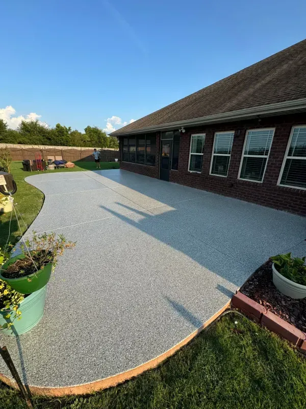 Curved gray patio beside a brick house, with potted plants and a lawn under a clear blue sky.