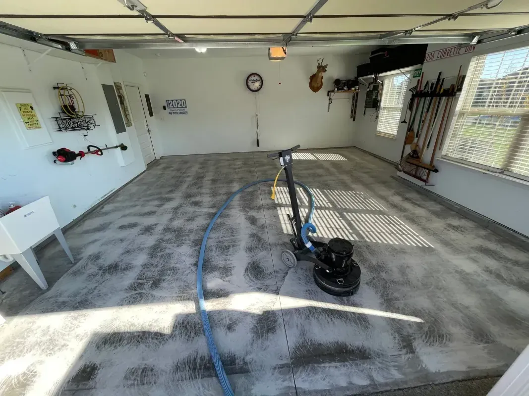 Empty garage with a pressure washer, hose, and streaked concrete floor drying in sunlight.