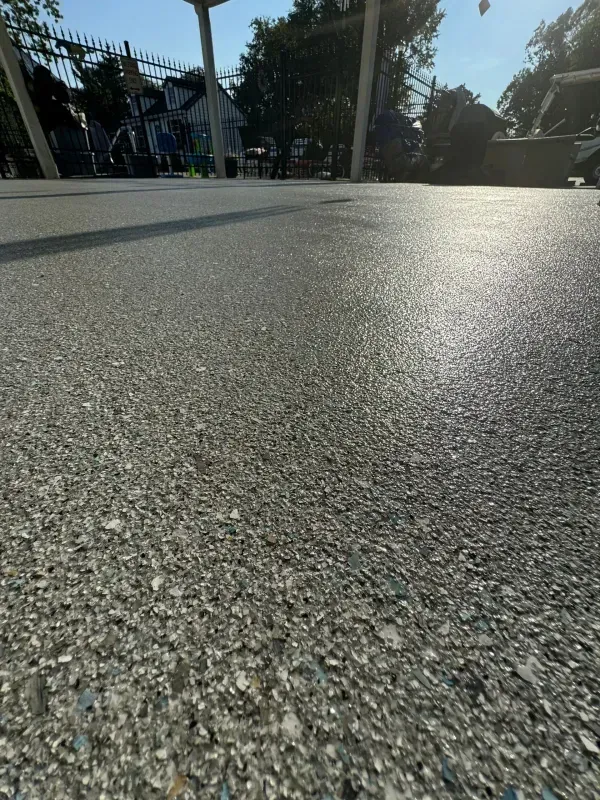 Close-up of textured gray pavement in bright sunlight with trees and buildings in the background