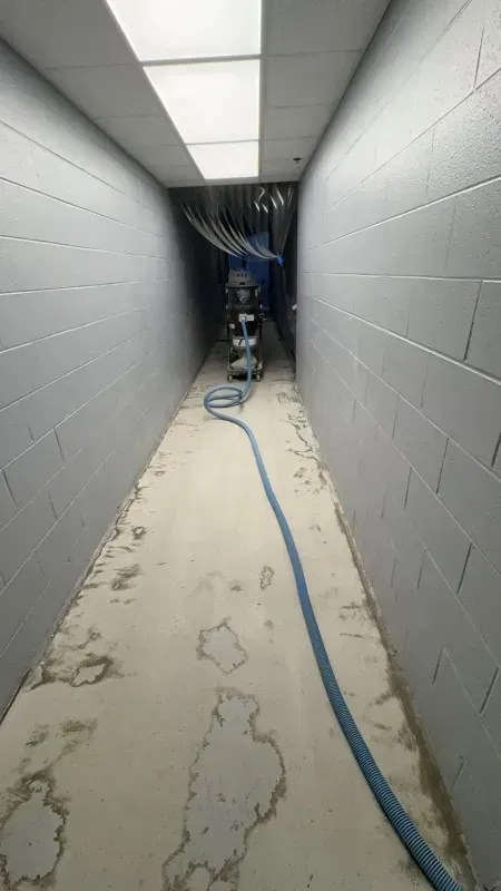 Narrow utility hallway with cinderblock walls, fluorescent lights, and a blue hose running along the floor