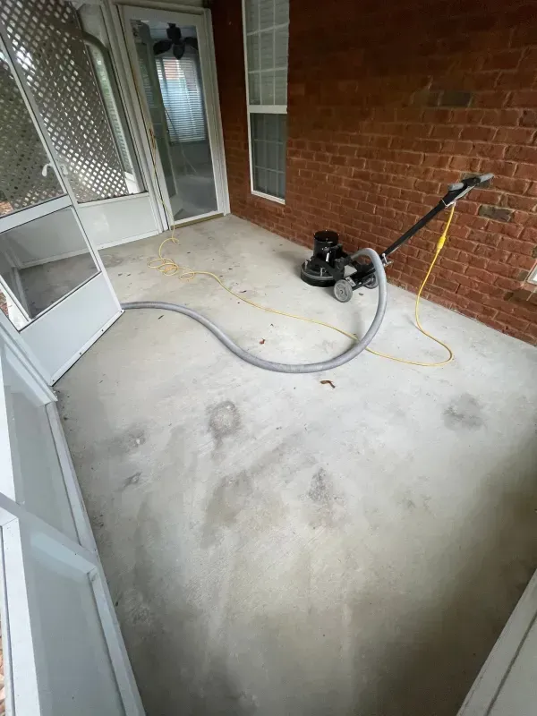 Porch floor being pressure-washed, with hose and cleaning machine near a brick wall.