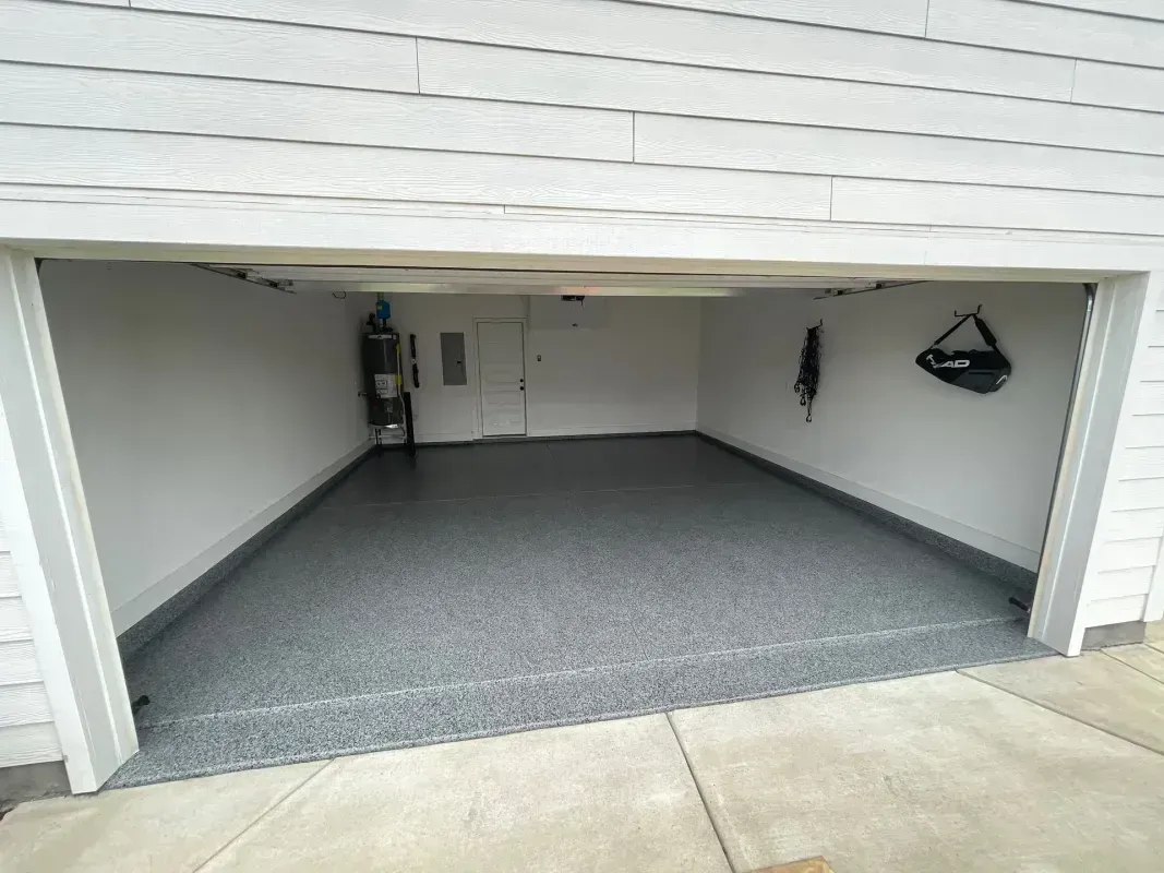 Empty garage with gray speckled floor, white walls, and the door open to the driveway