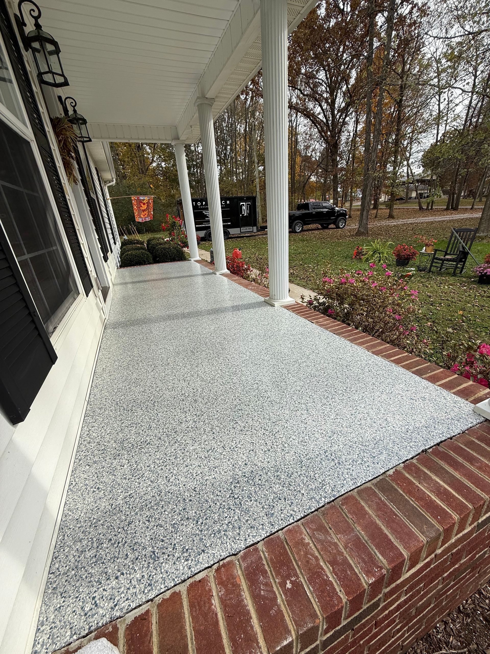 Front porch with white railing, brick step, and speckled gray flooring beside a wooded yard.