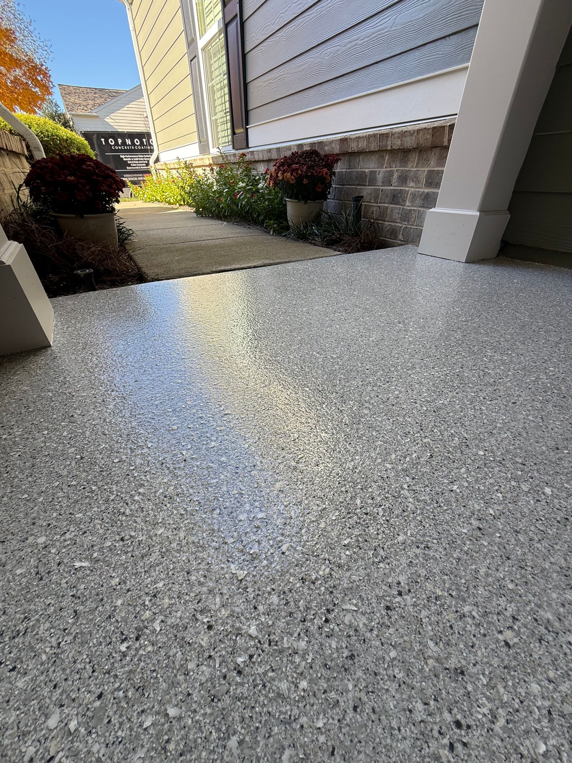 Concrete patio beside a house with shrubs and a sunlit walkway leading toward a side yard