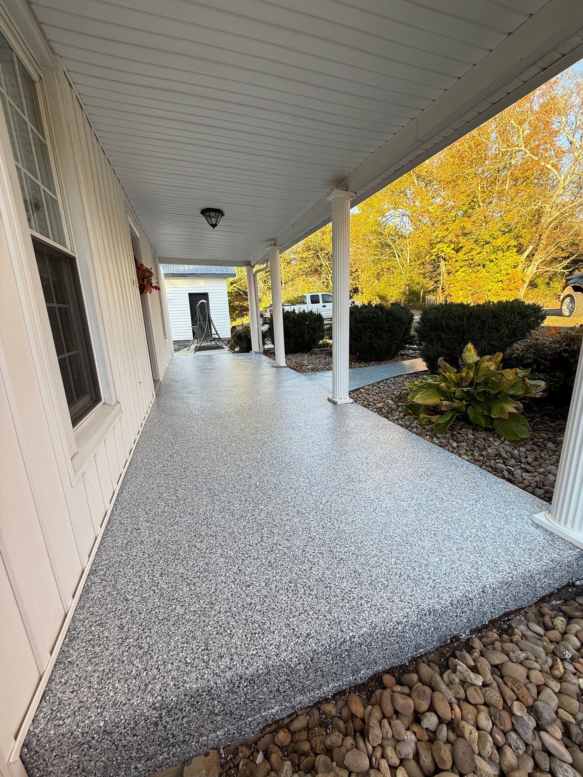 Covered porch with white columns, speckled gray floor, and fall shrubs along the side of a house.