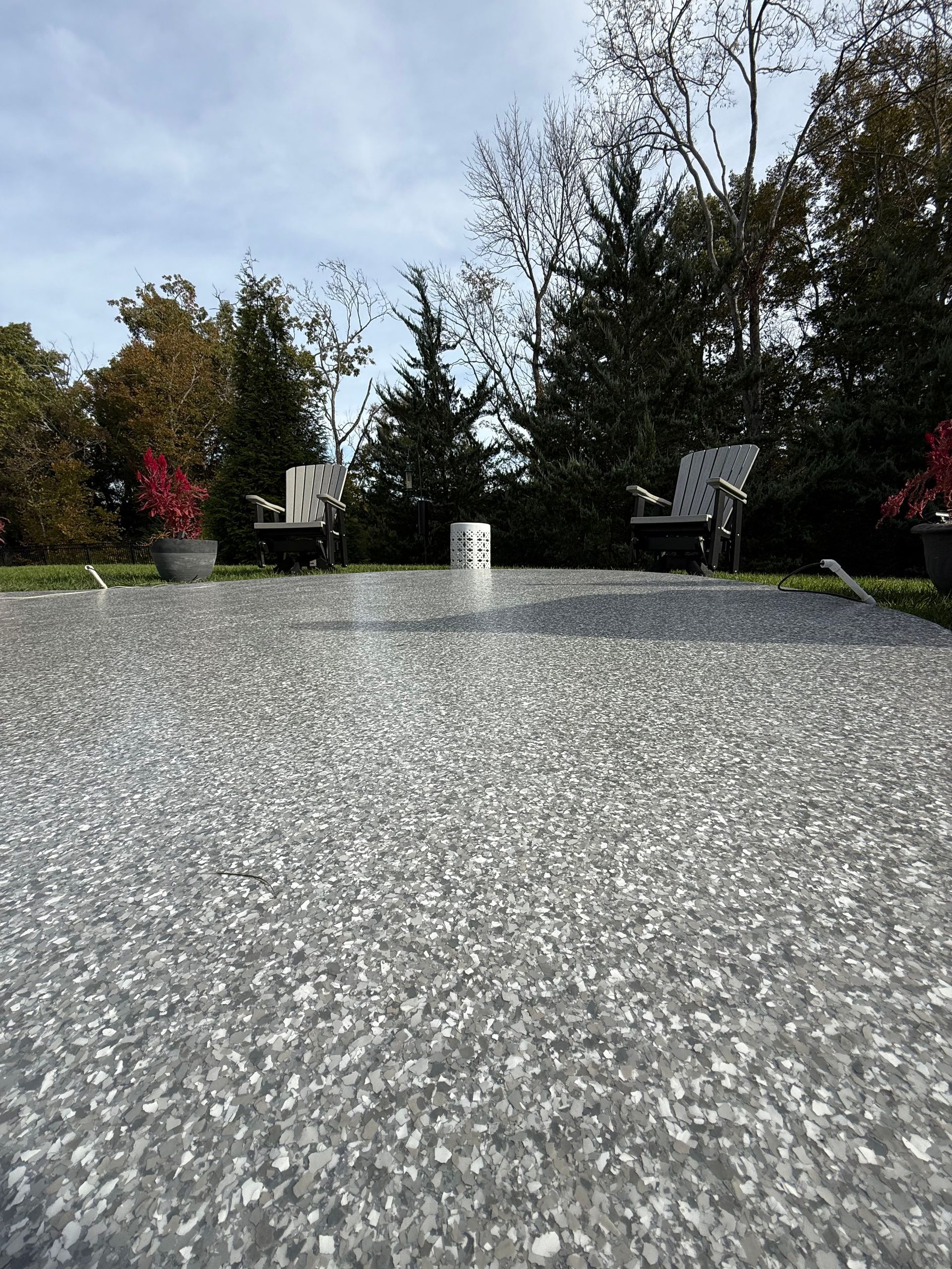 Backyard patio with speckled stone surface, chairs, potted flowers, and trees under a cloudy sky
