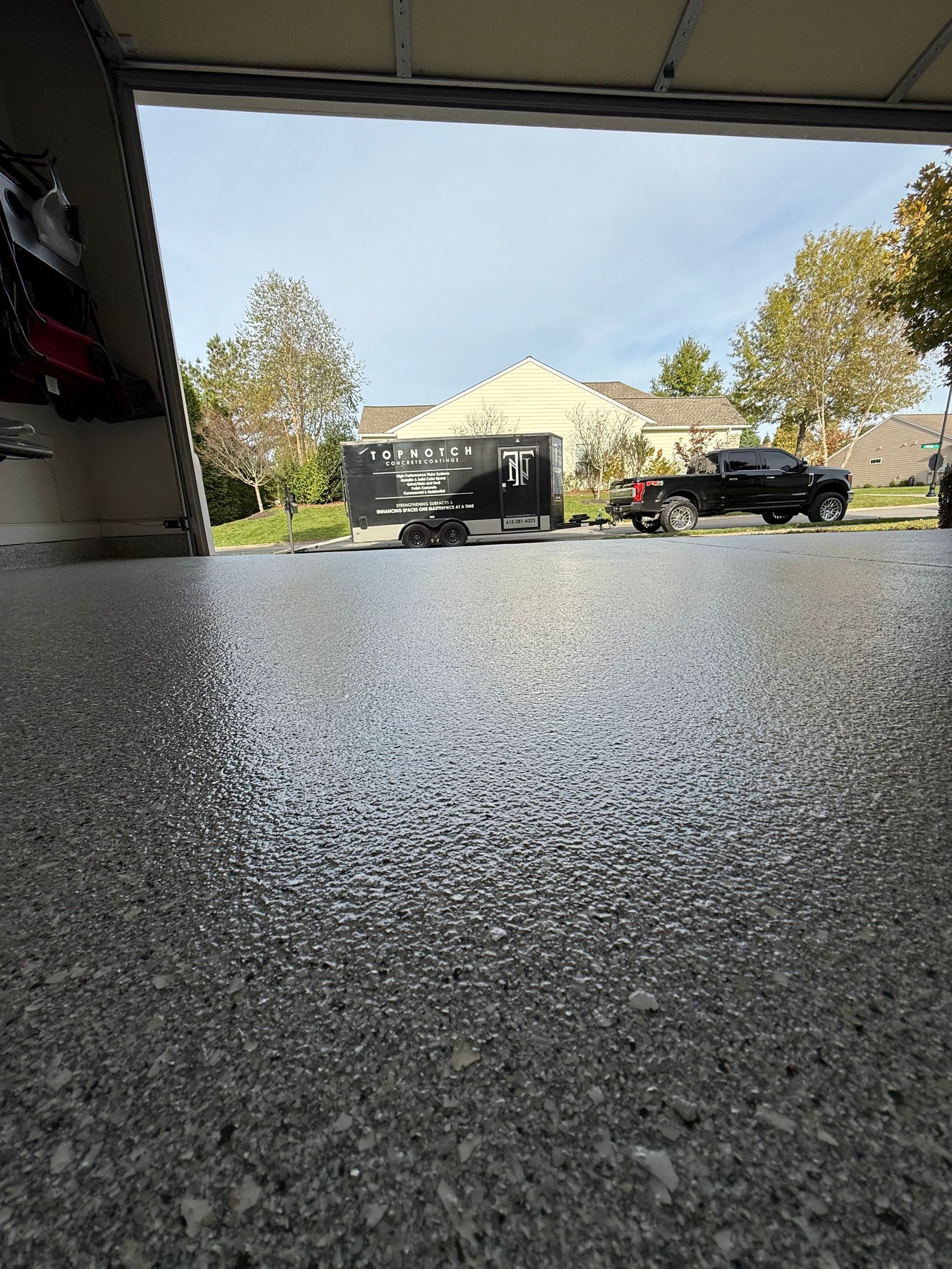 Driveway view from a garage floor toward a suburban street with a house, trees, and parked vehicles.