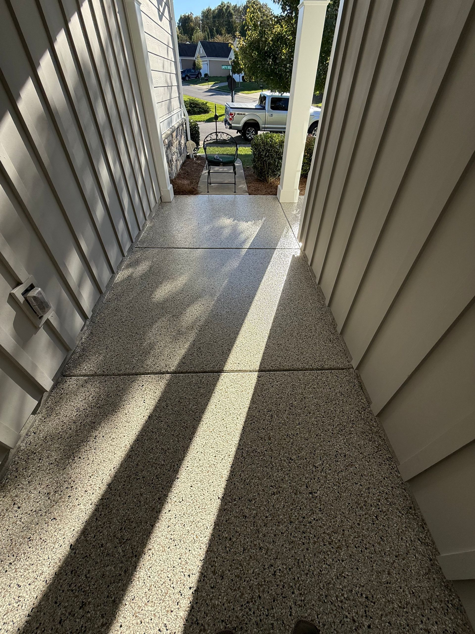 Narrow sunlit concrete walkway between beige siding, leading to a porch and street below.