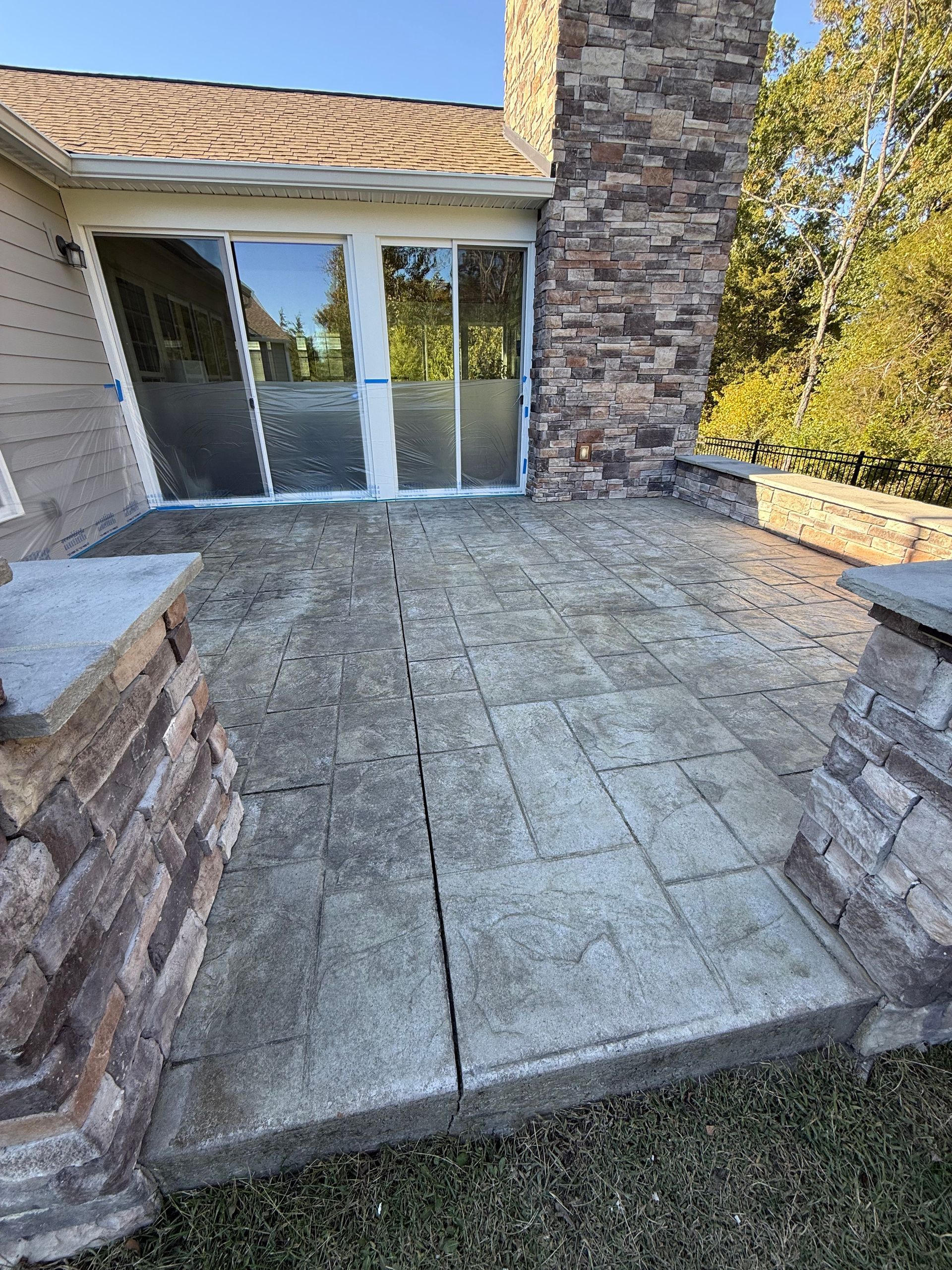 Stone patio with cracked pavers beside a house, leading to sliding glass doors and a stone column.