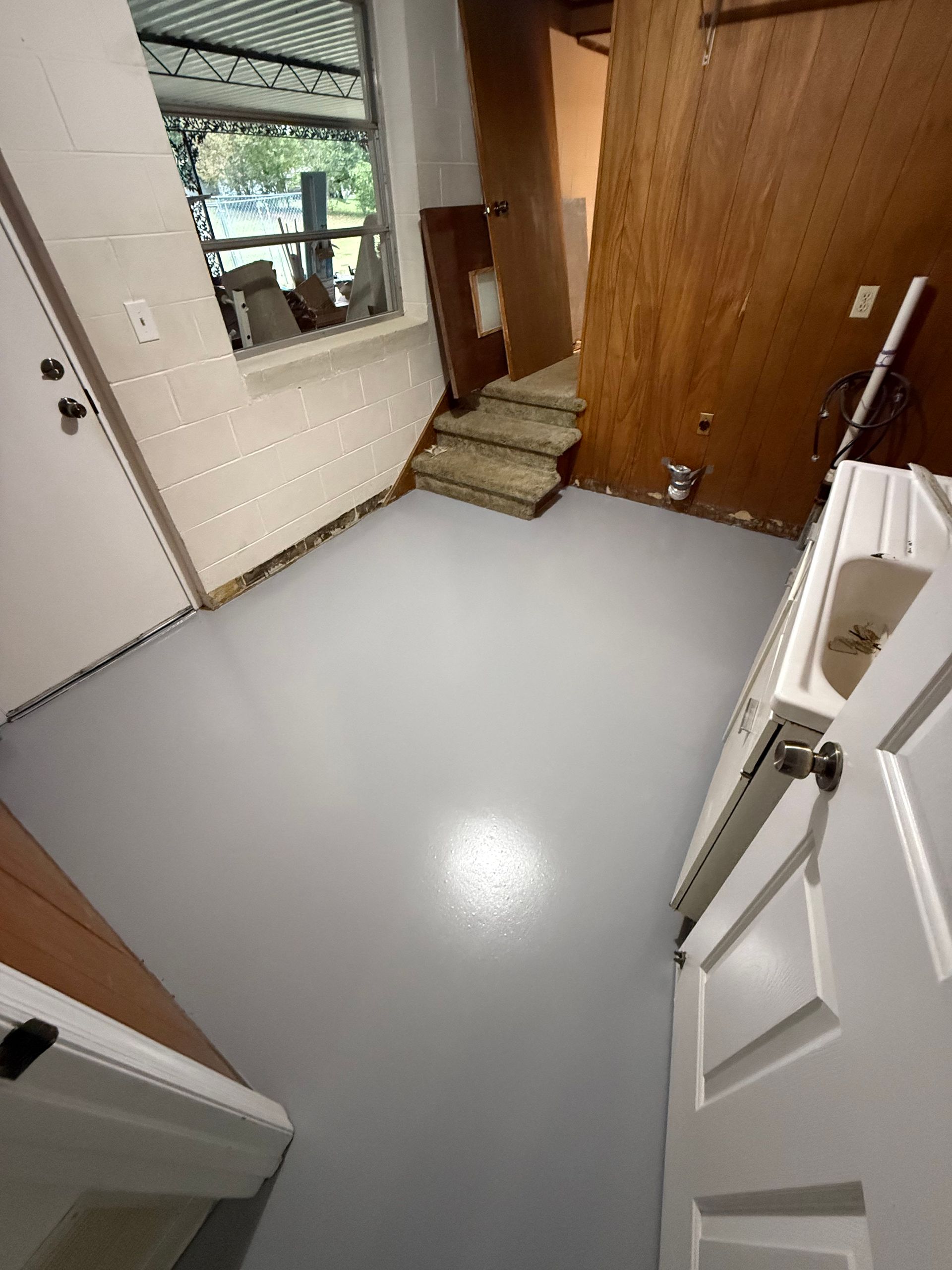 Empty room with gray floor, white door, stairs, and wood-paneled wall.