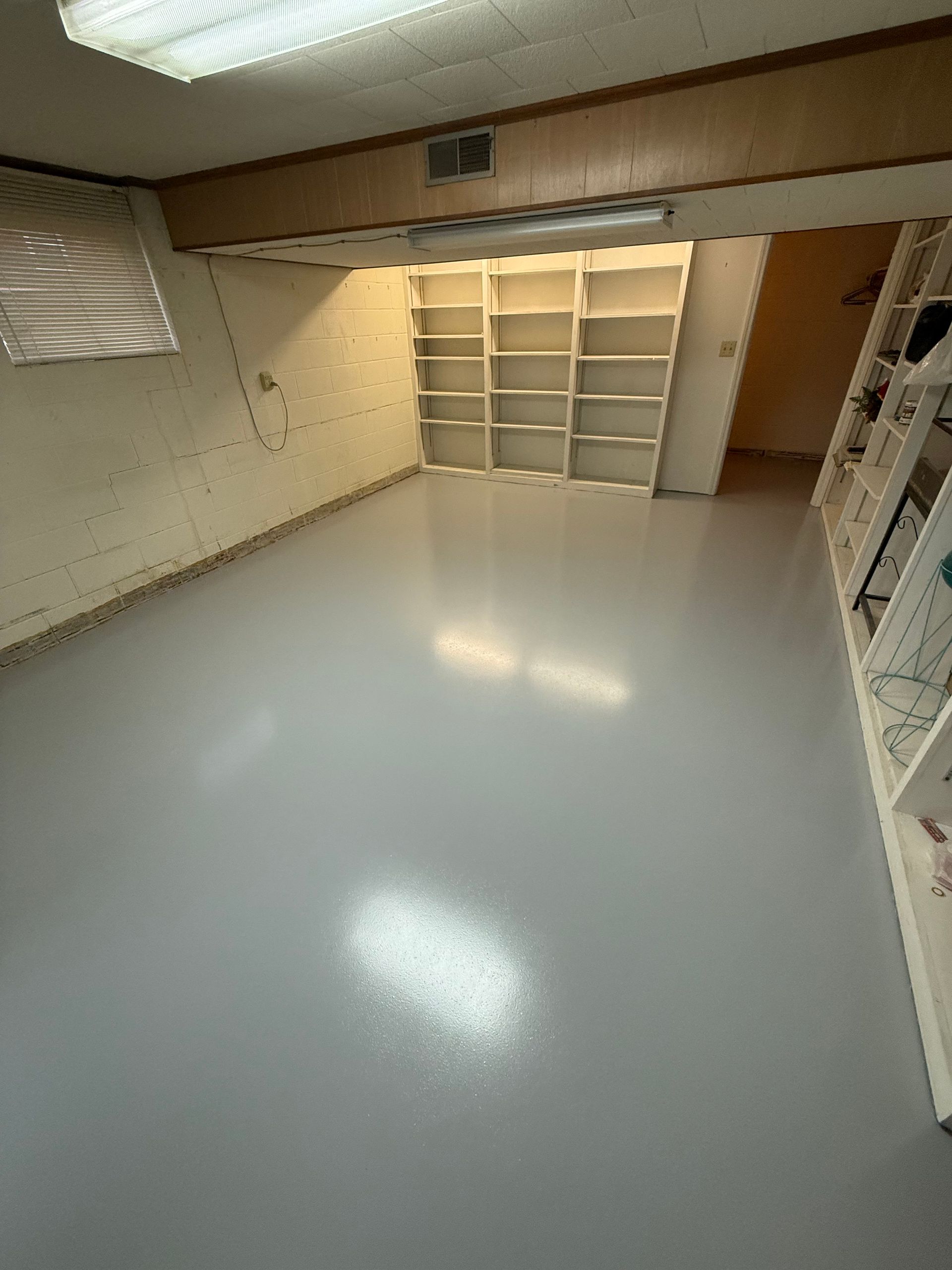 Empty basement room with glossy gray floor, beige walls, and built-in shelving along the back wall