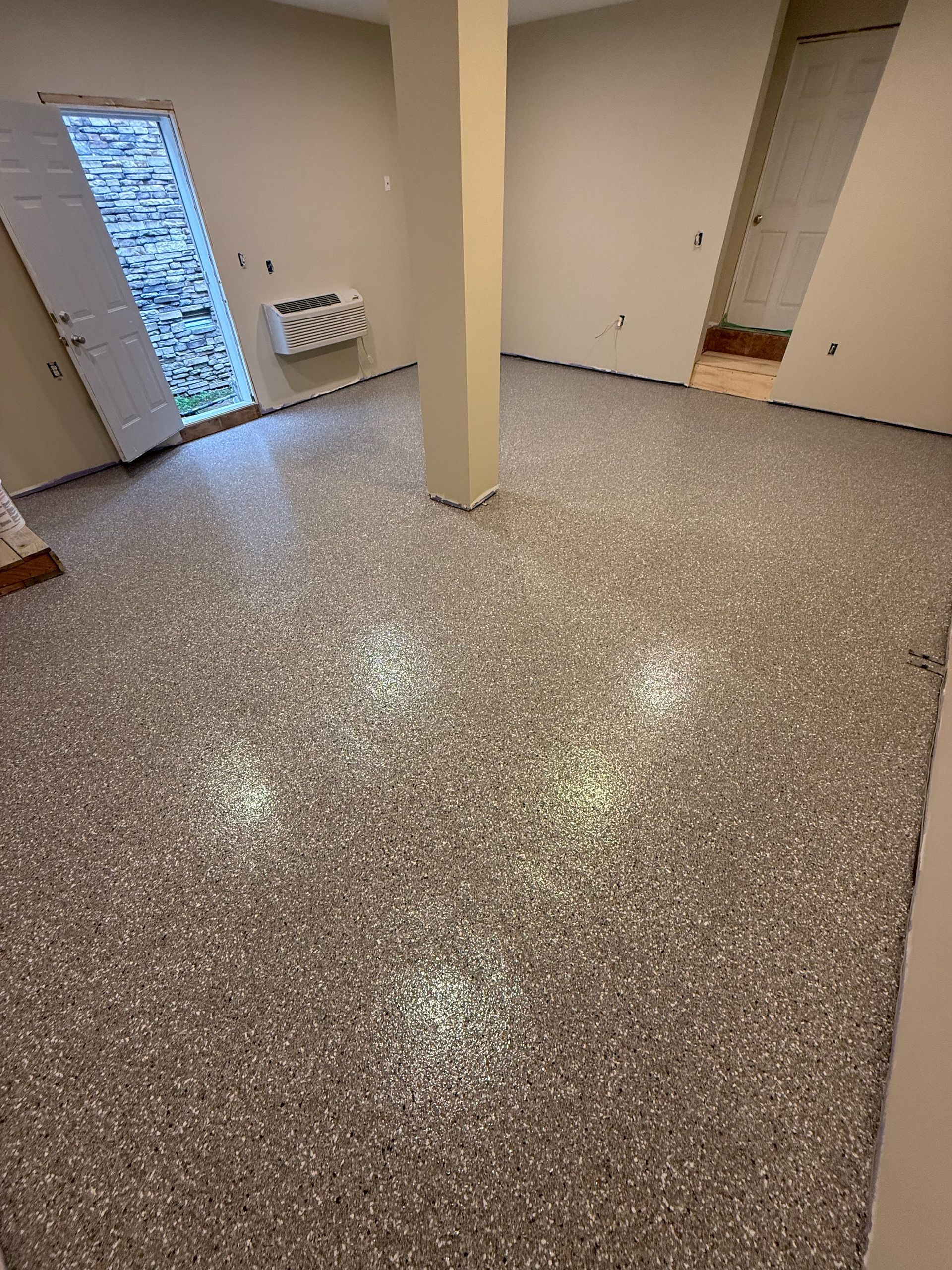 Empty beige room with speckled floor, central pillar, and windows on the left.