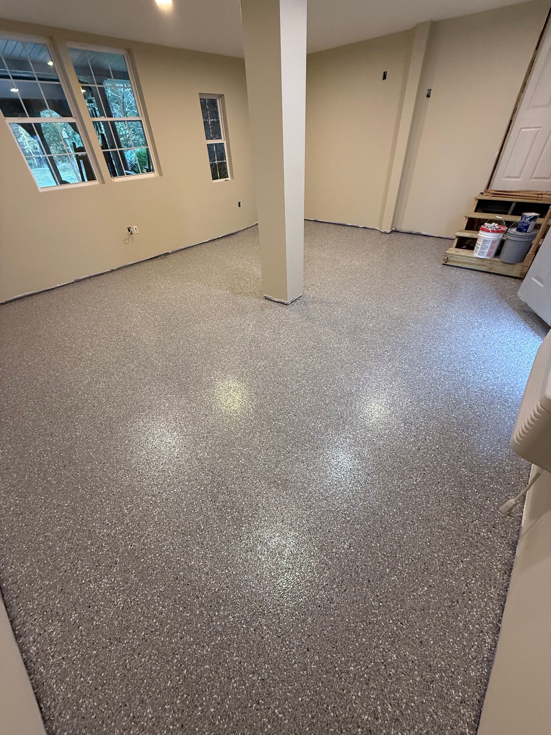 Empty basement room with gray speckled floor, white walls, windows, and a support column