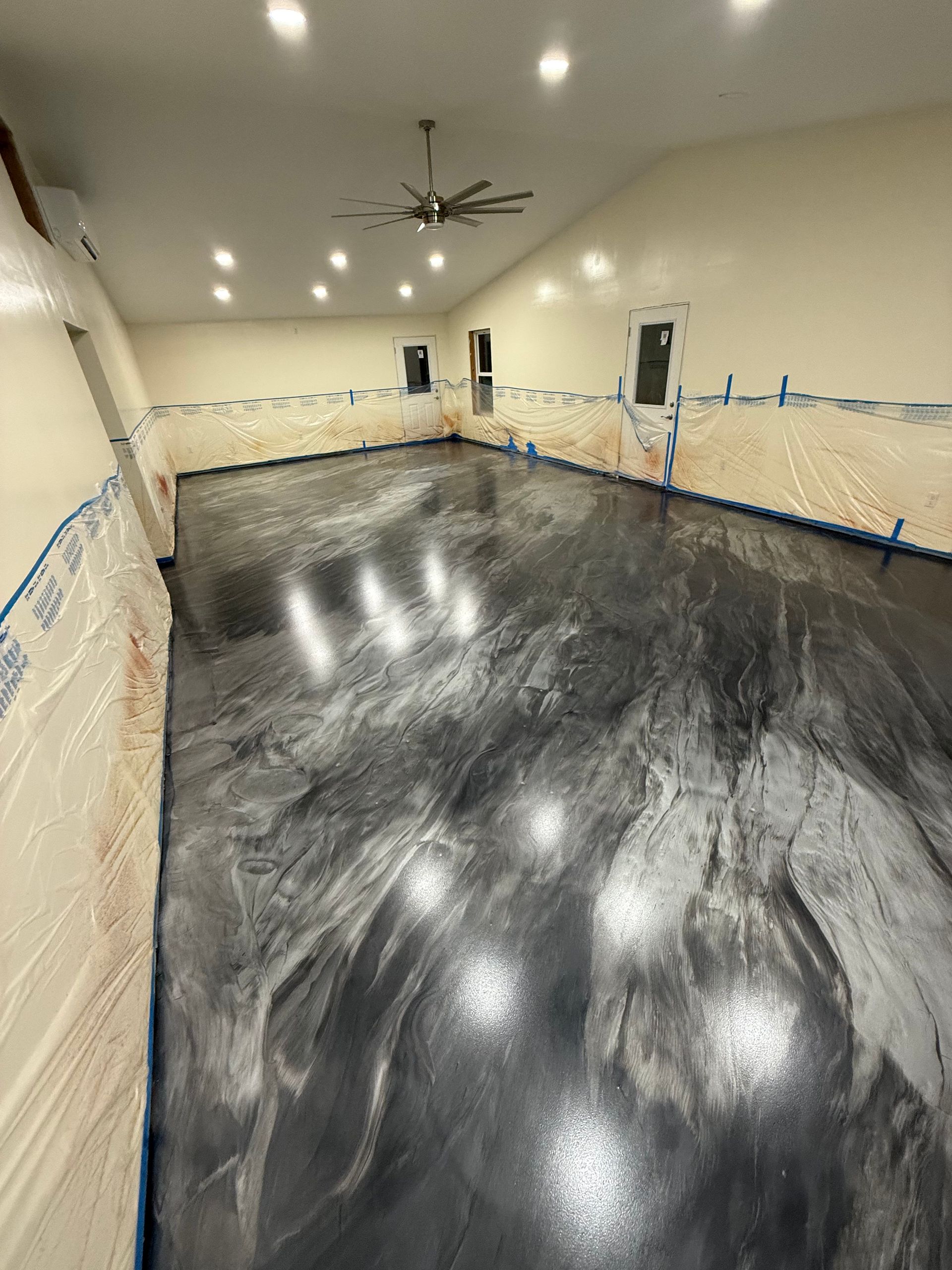 Empty room with glossy black epoxy floor, beige walls, ceiling fan, and blue painter’s tape along the baseboards