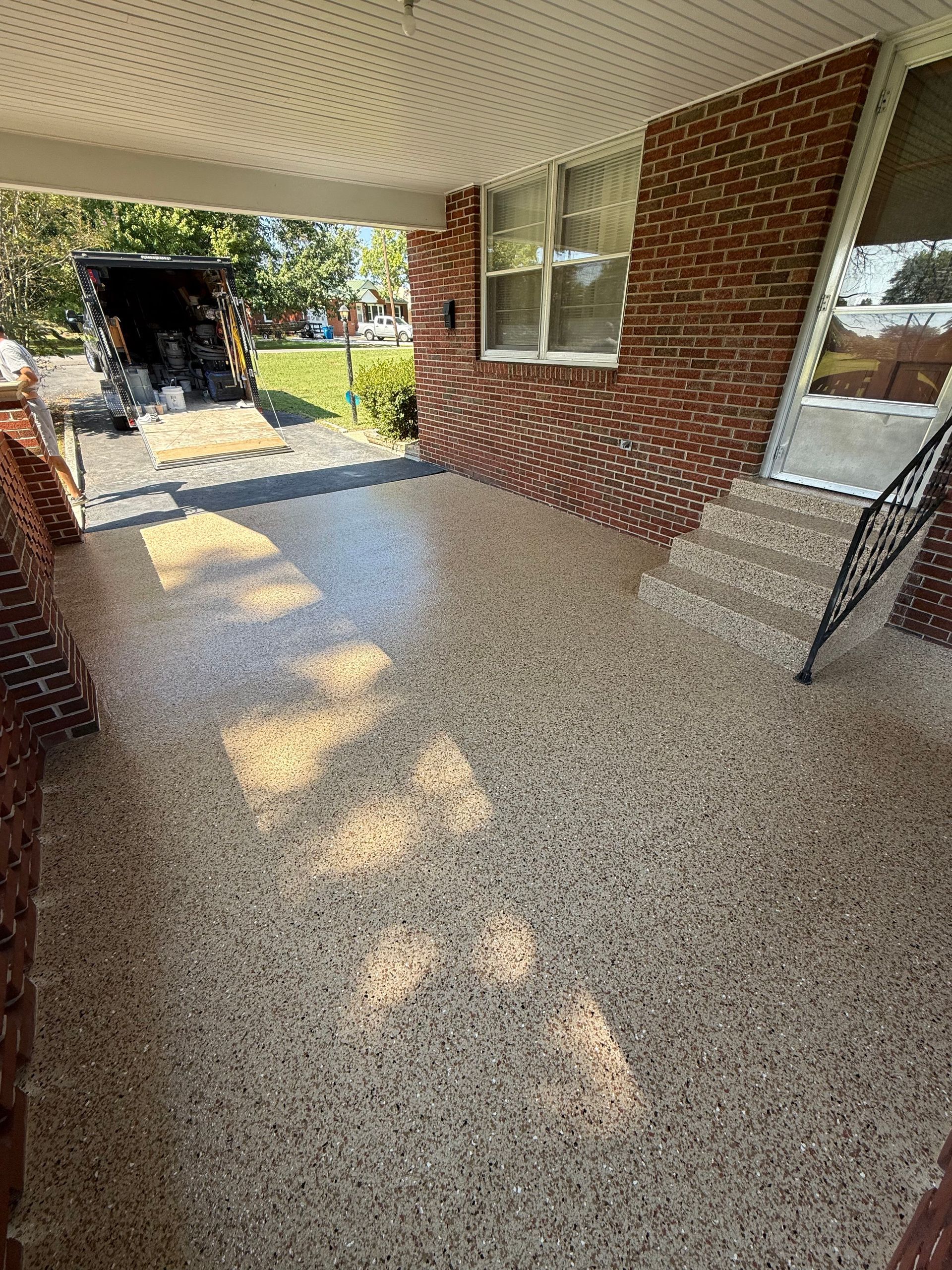 Covered outdoor patio with speckled flooring, brick railing, and trees beyond the open side
