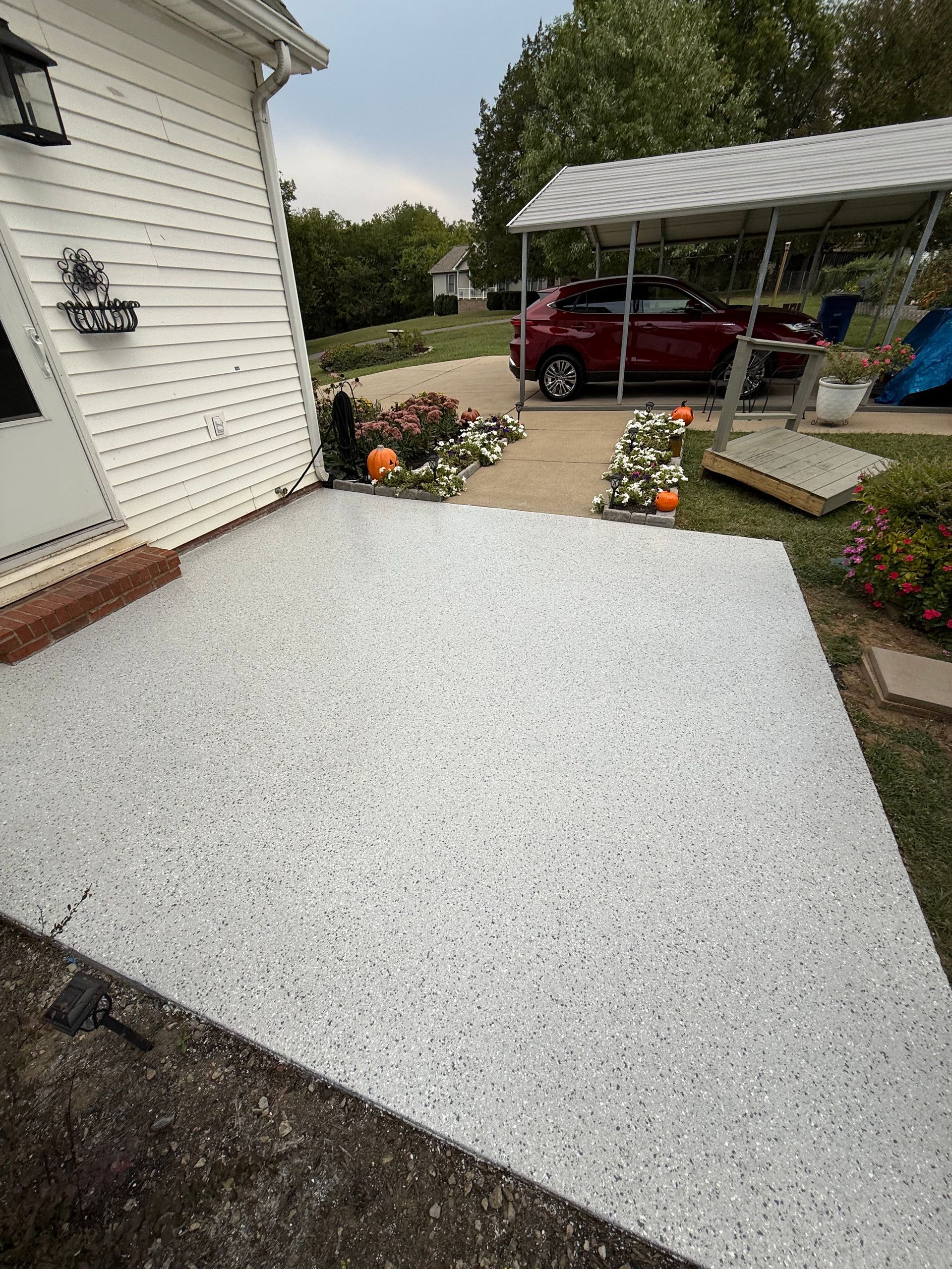 White-speckled concrete patio beside a house, with flower beds and a red SUV under a carport.