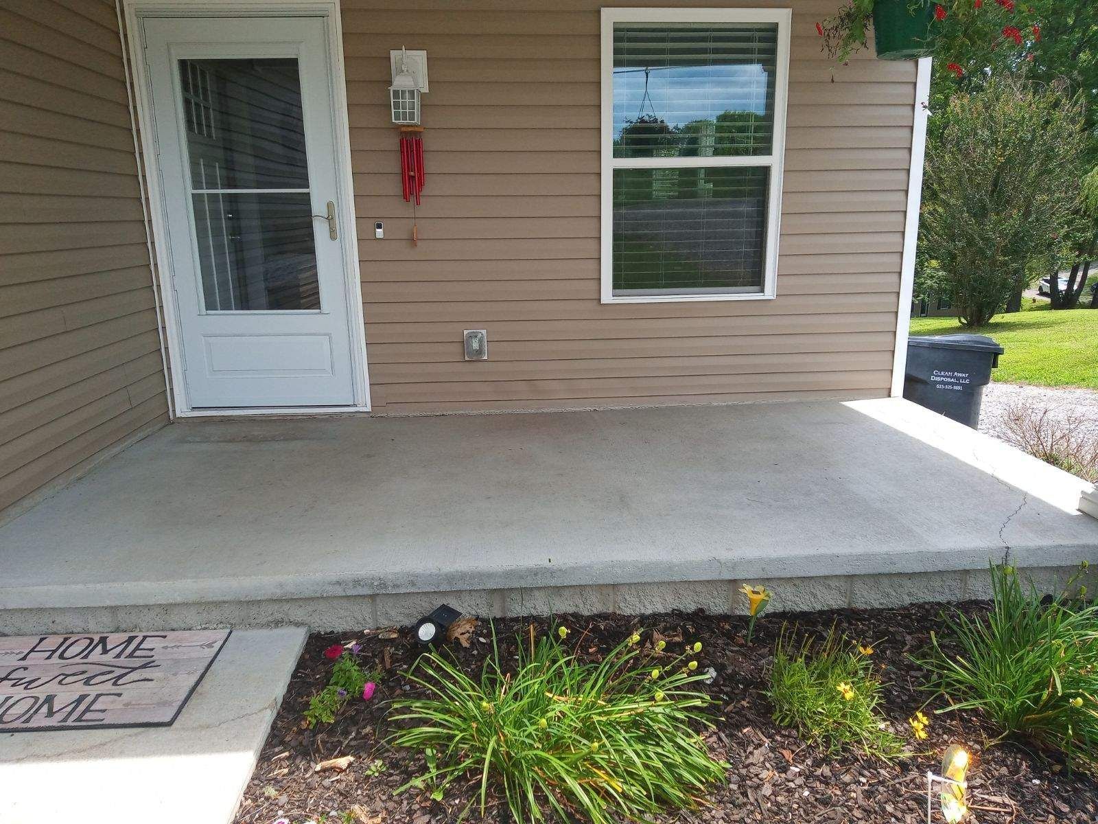 A tan-sided house with a concrete porch, white door, window, and a small flower bed.
