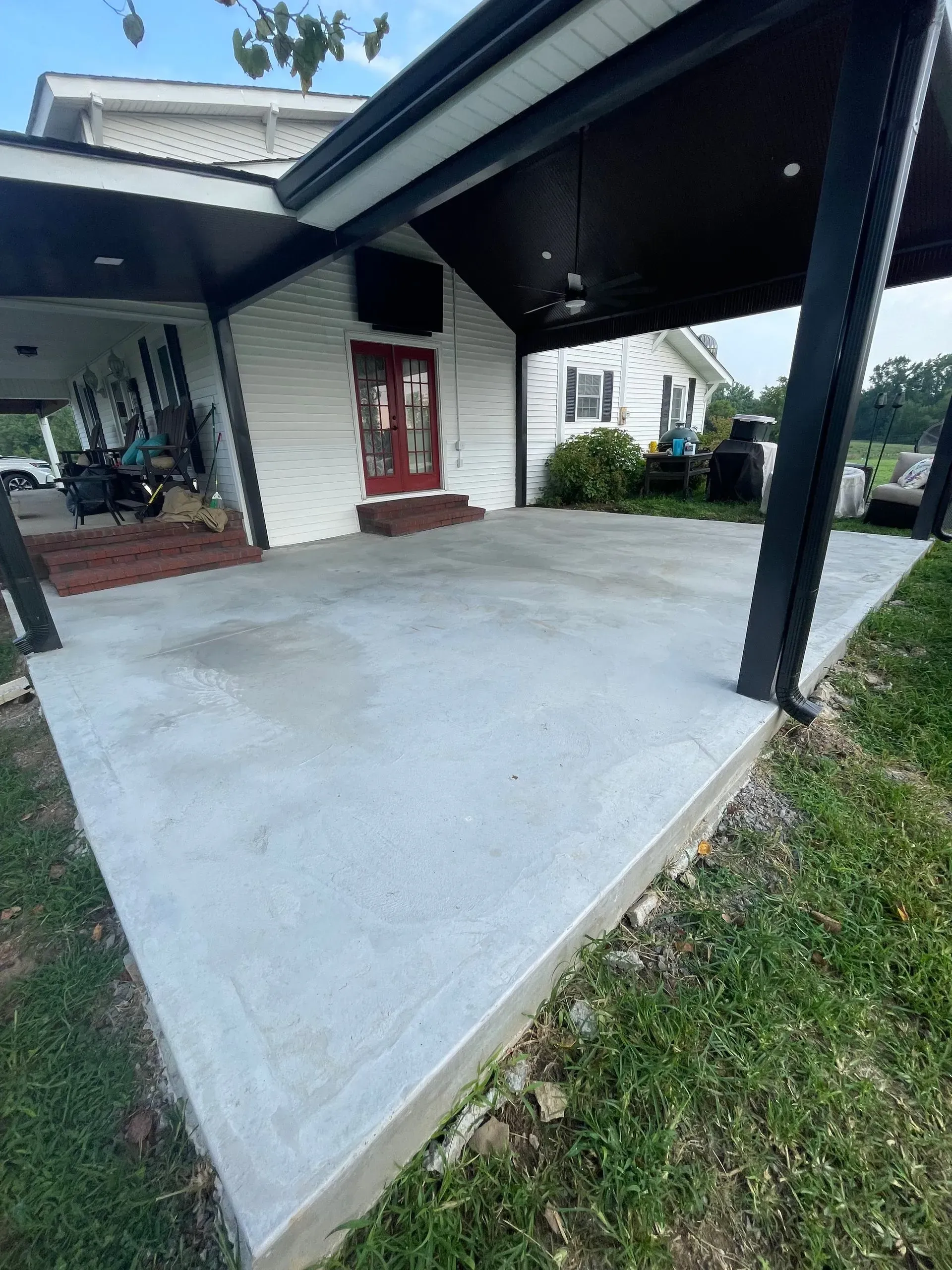 Concrete patio under a black-trimmed overhang next to a white house with red doors.