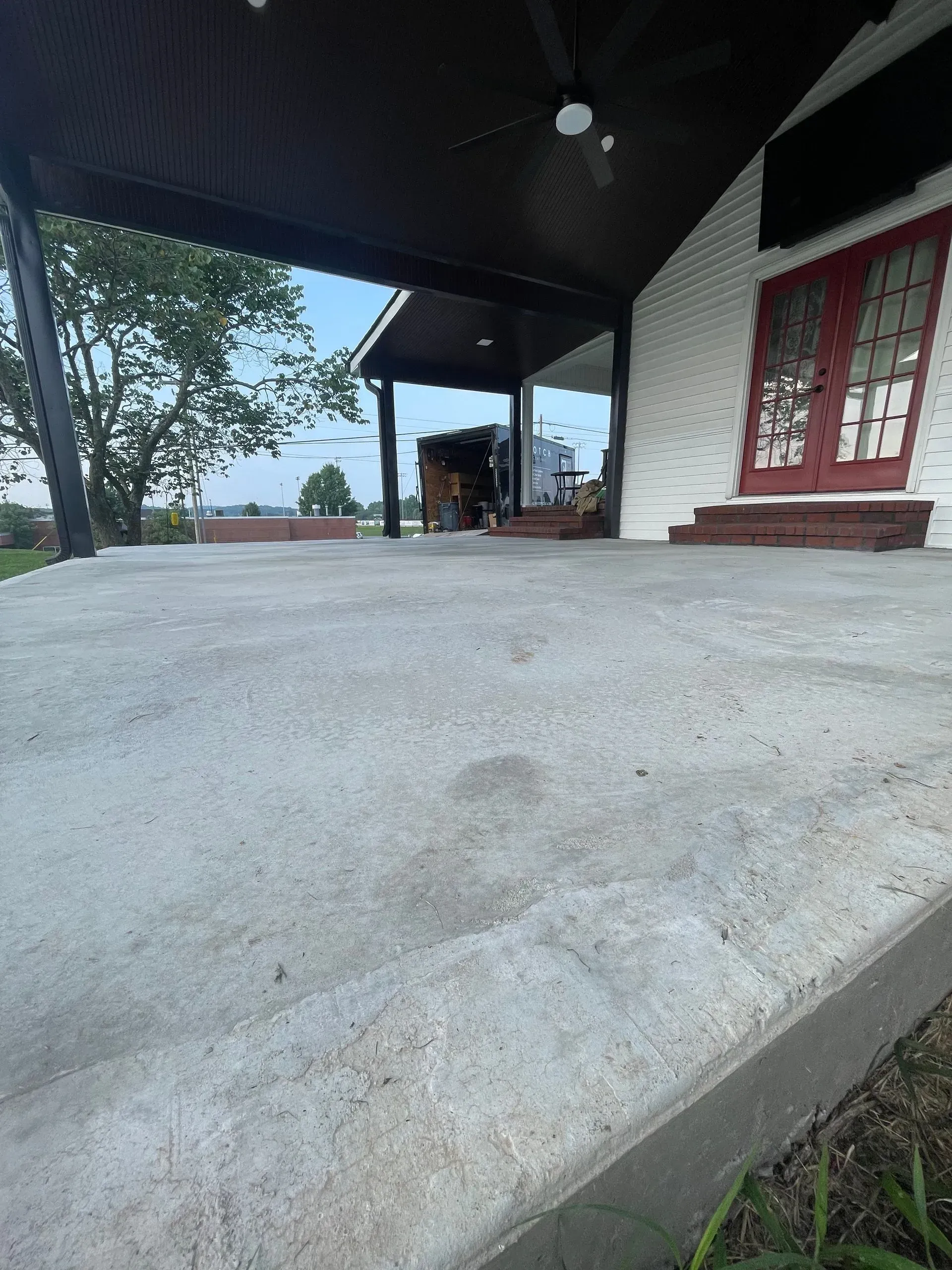 A concrete porch of a white brick building with red doors and dark overhang; grass in foreground, trees visible in background.