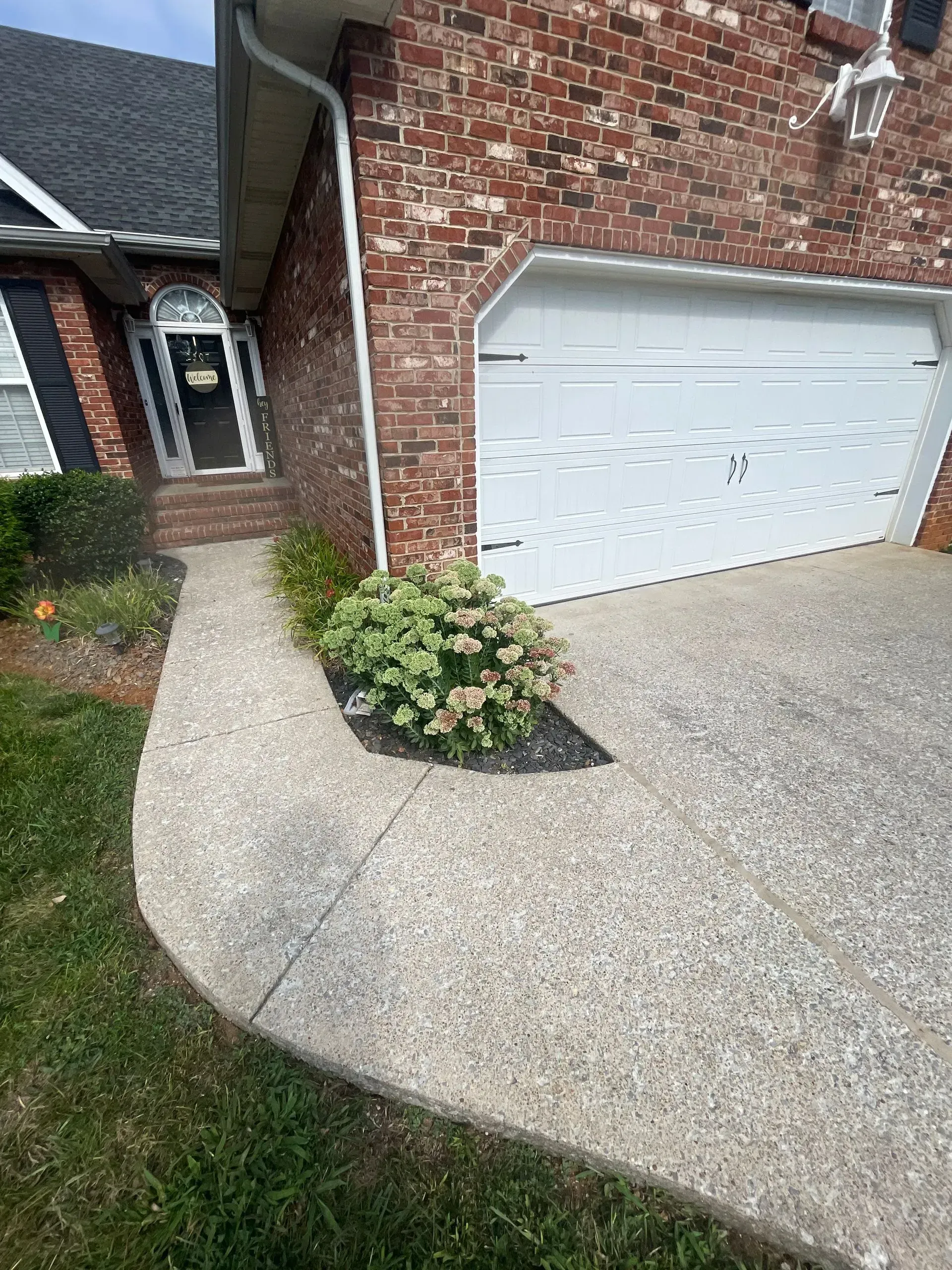 Concrete path leading to a brick house with a white garage door and shrubbery.