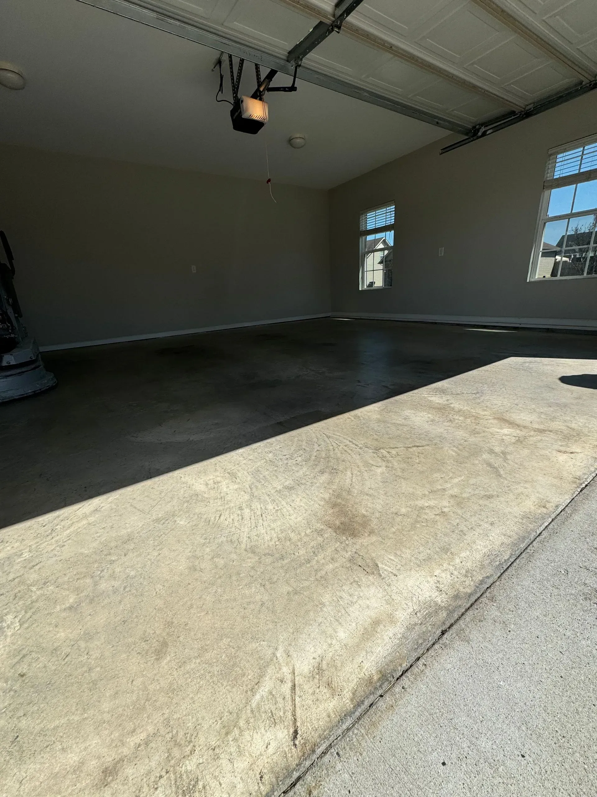 Empty garage with grey concrete floor, walls, and open garage door. Two windows, sunlight, empty space.