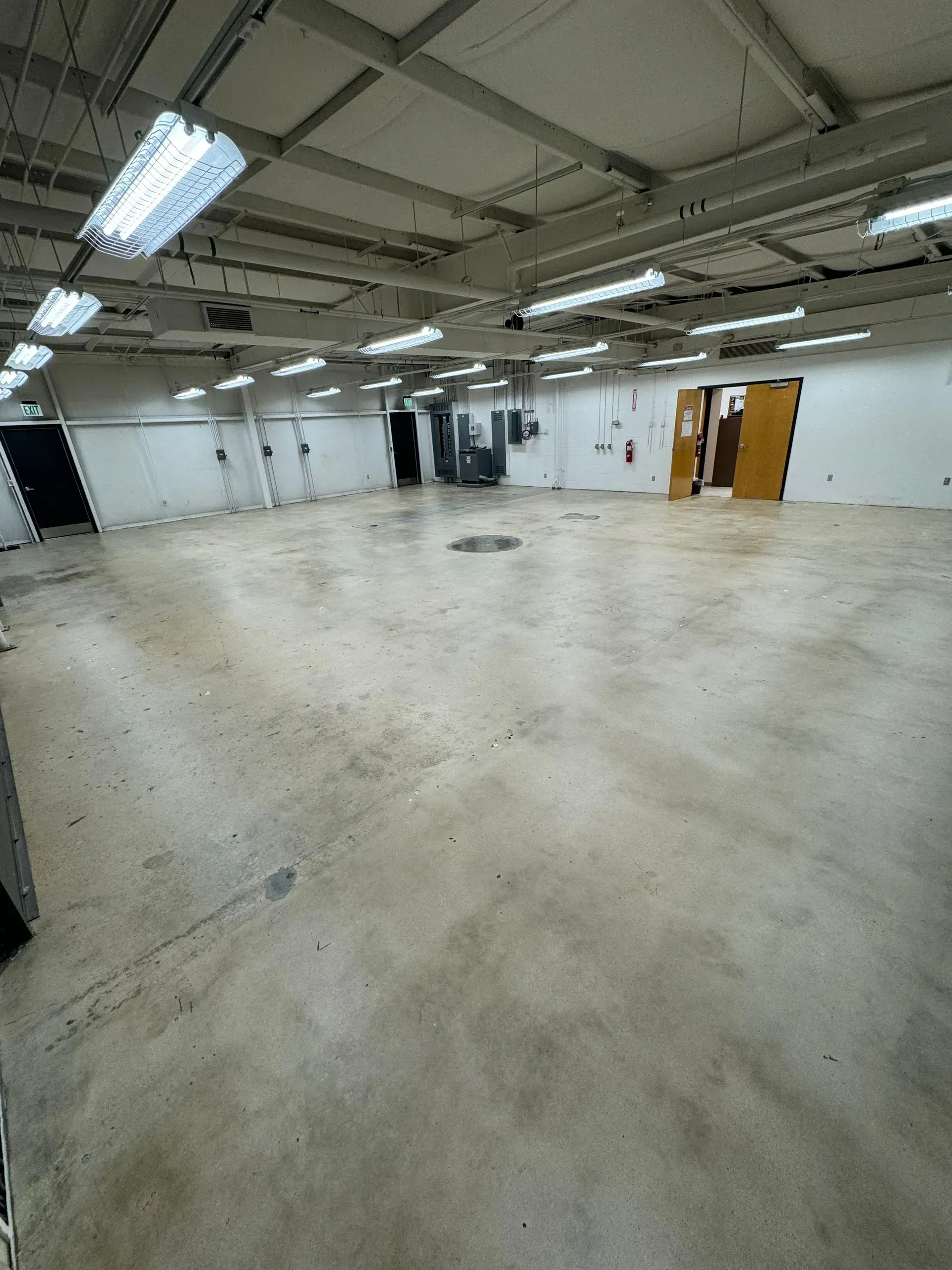 Empty, large warehouse with concrete floor, white walls, and fluorescent lights.  Doors visible in the distance.